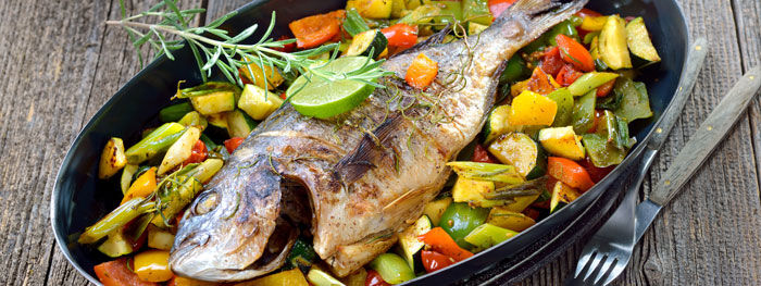 Gegrillter Fisch mit Gemüse, Rosmarin und Limette im ovalen Teller auf Holzhintergrund